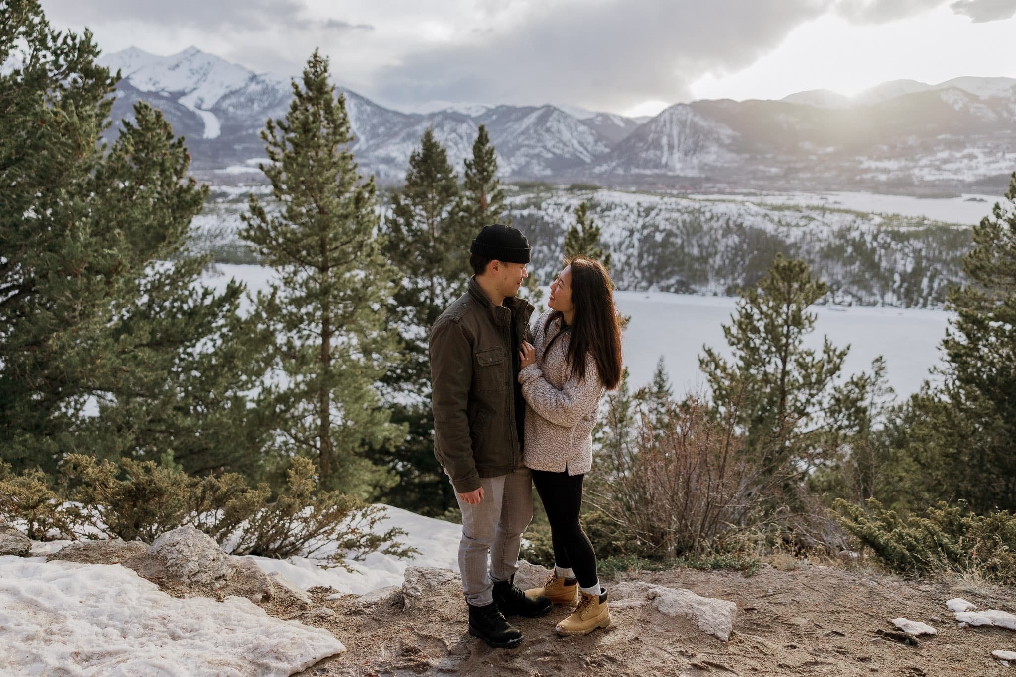 Couple standing among pines on a snowy overlook above a mountain lake